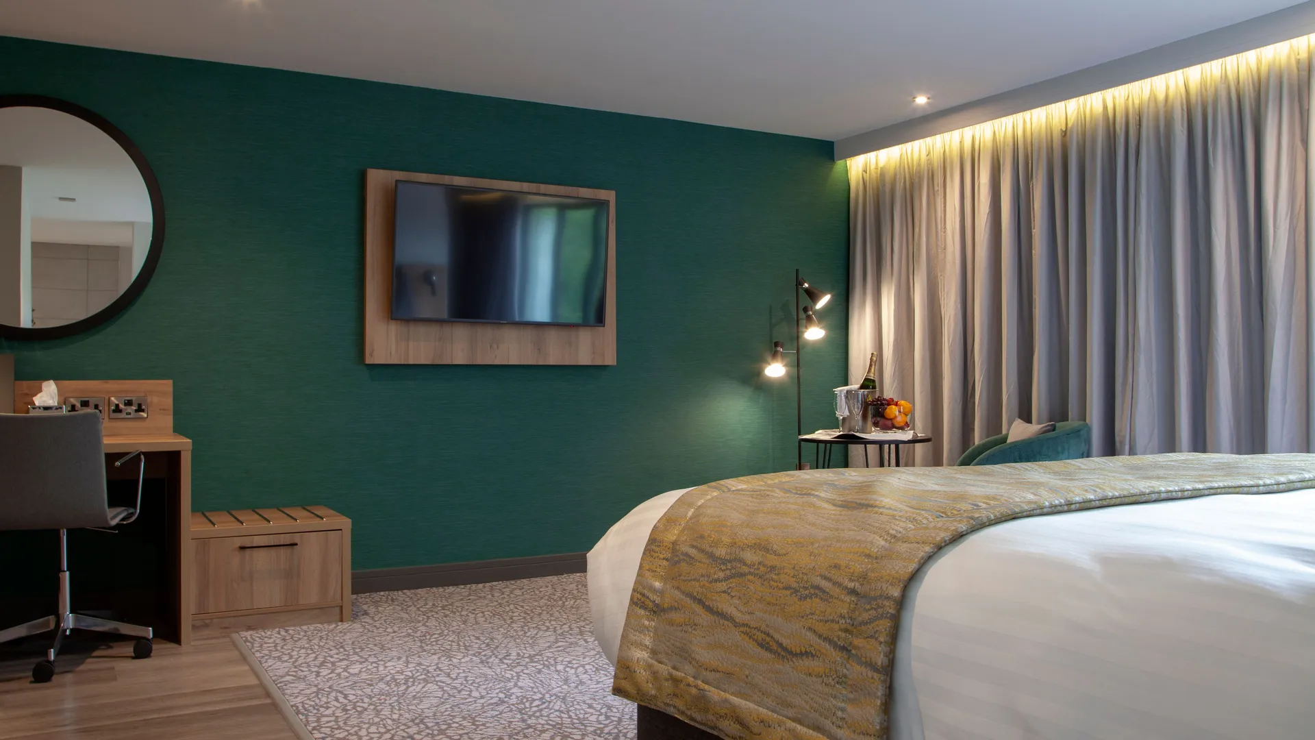 Modern hotel bedroom with teal feature wall, king-size bed, desk and wall-mounted television.
