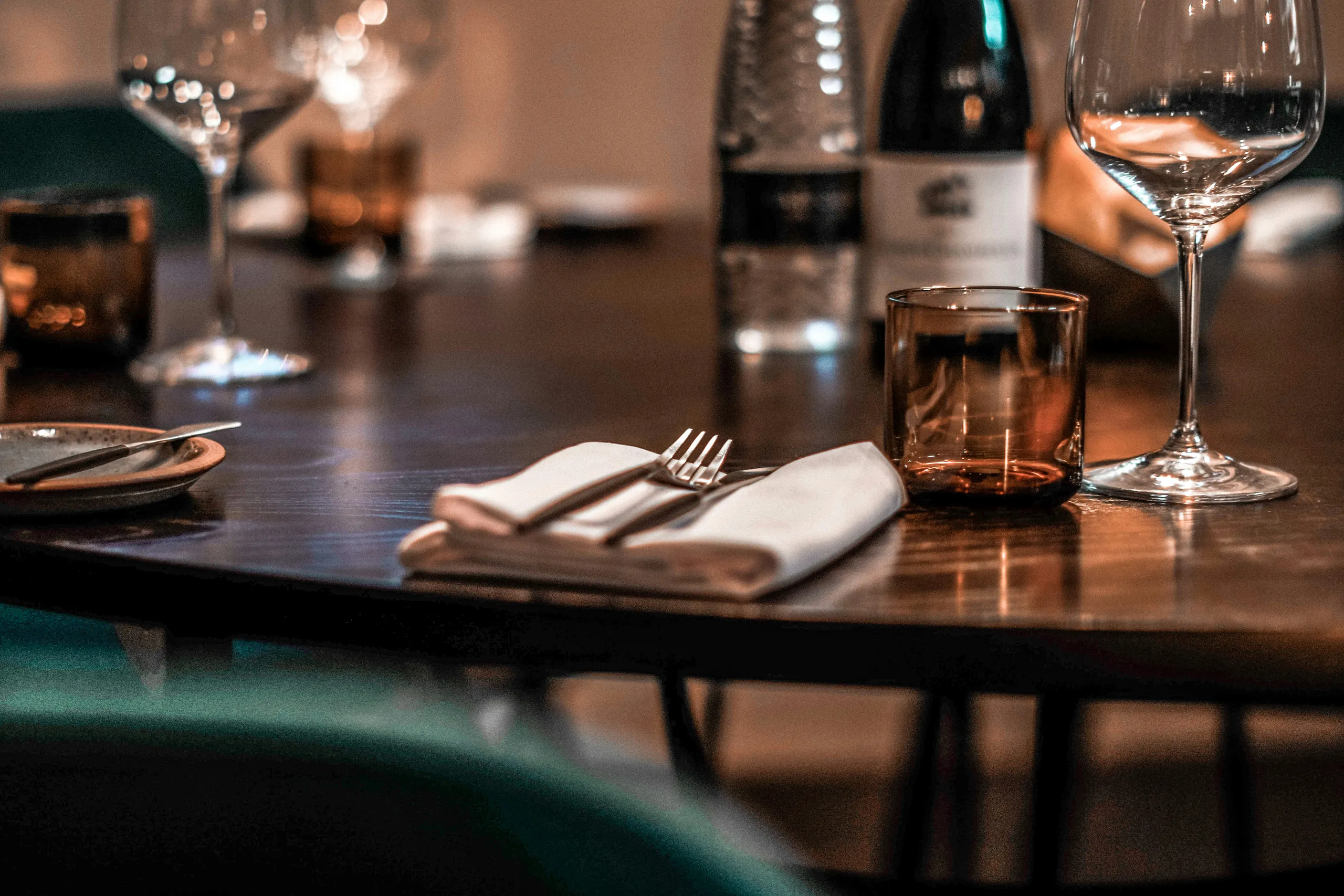 Warmly lit restaurant table set with wine glasses, a bottle, folded napkins and cutlery.