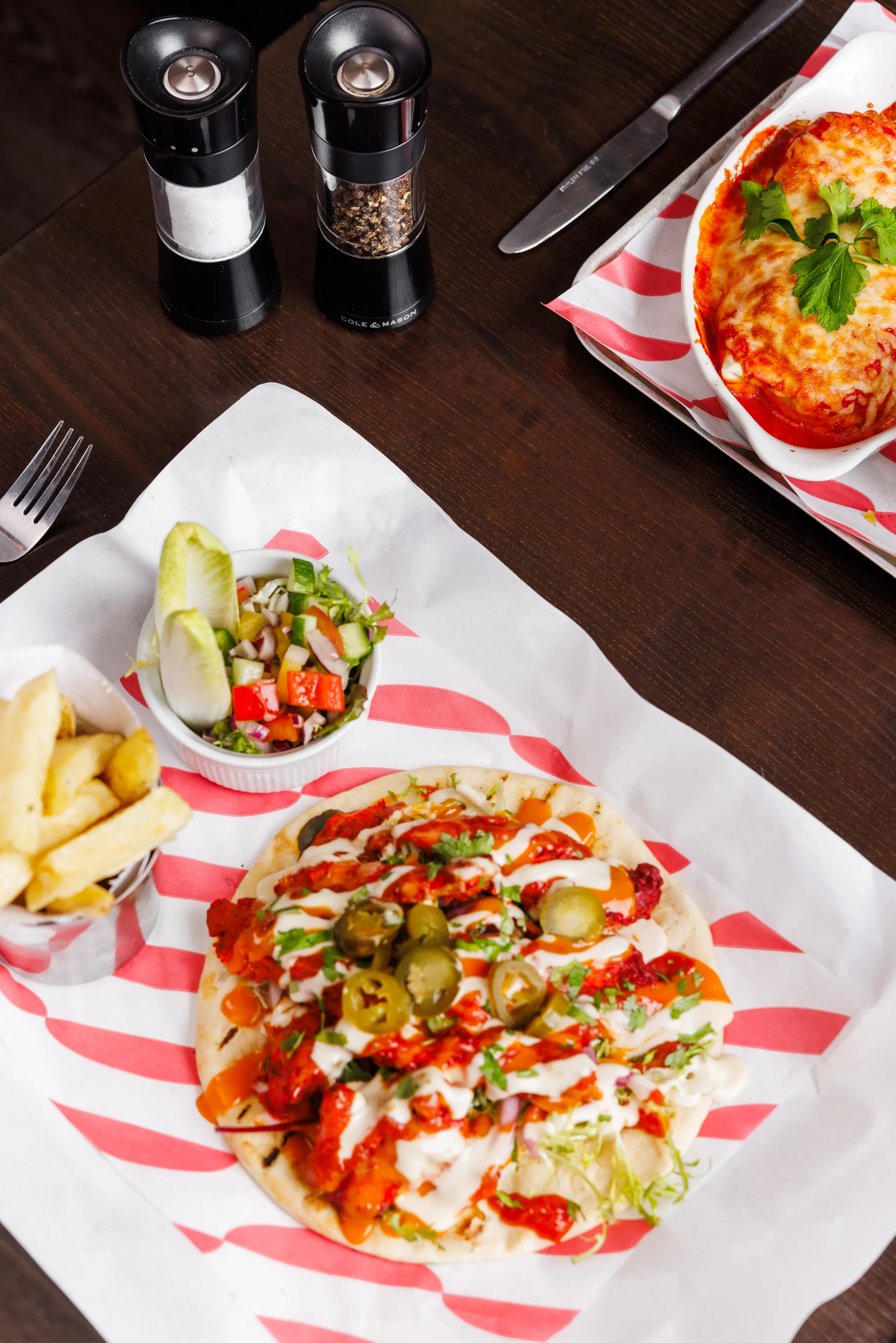 Loaded flatbread topped with jalapeños, sauces and herbs, served with chips, salad and a baked cheese dish on a wooden table.