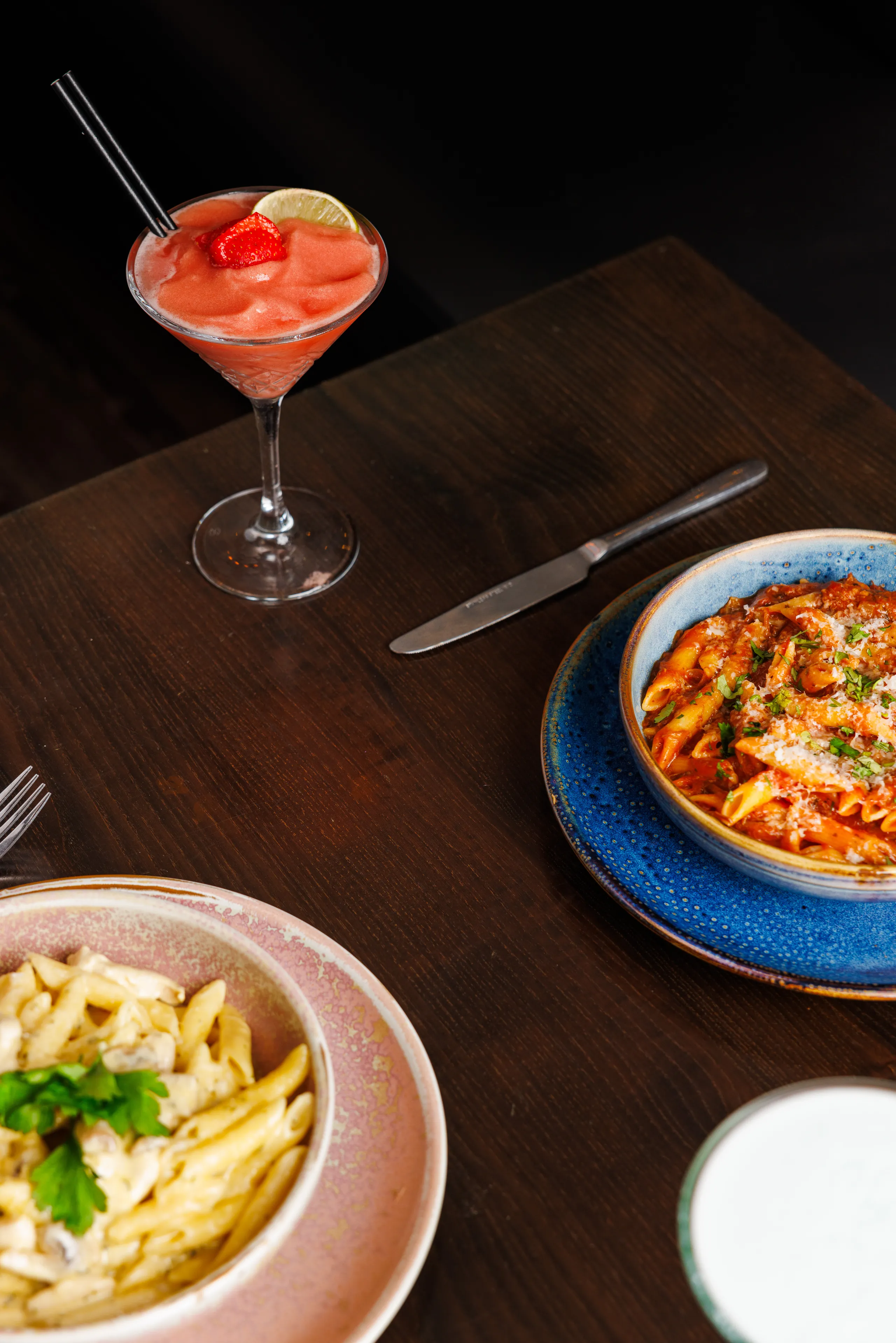 Strawberry frozen cocktail with lime beside bowls of pasta on a dark wooden table