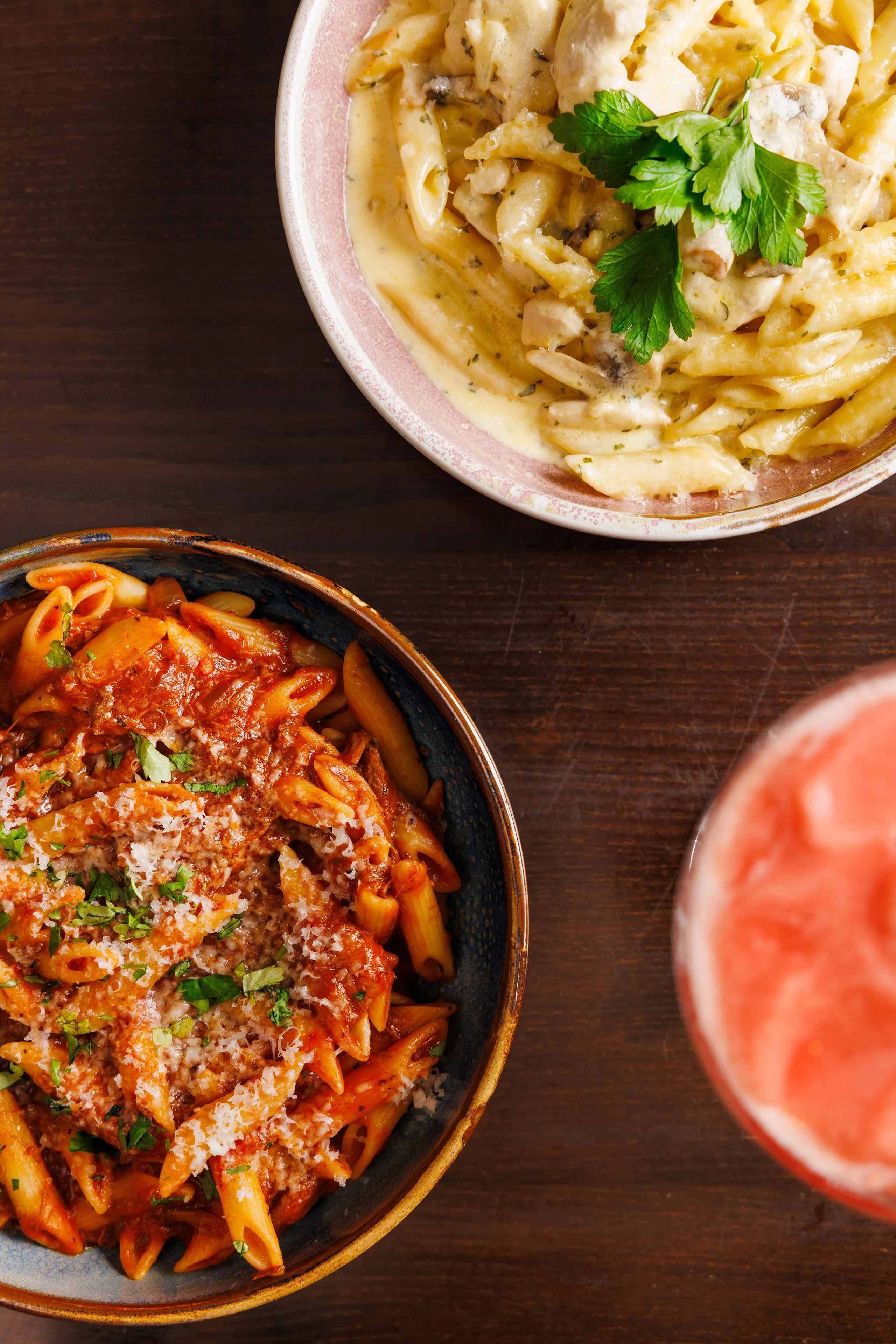 Two bowls of pasta on a dark wooden table, one creamy with parsley and one tomato meat sauce with grated cheese.