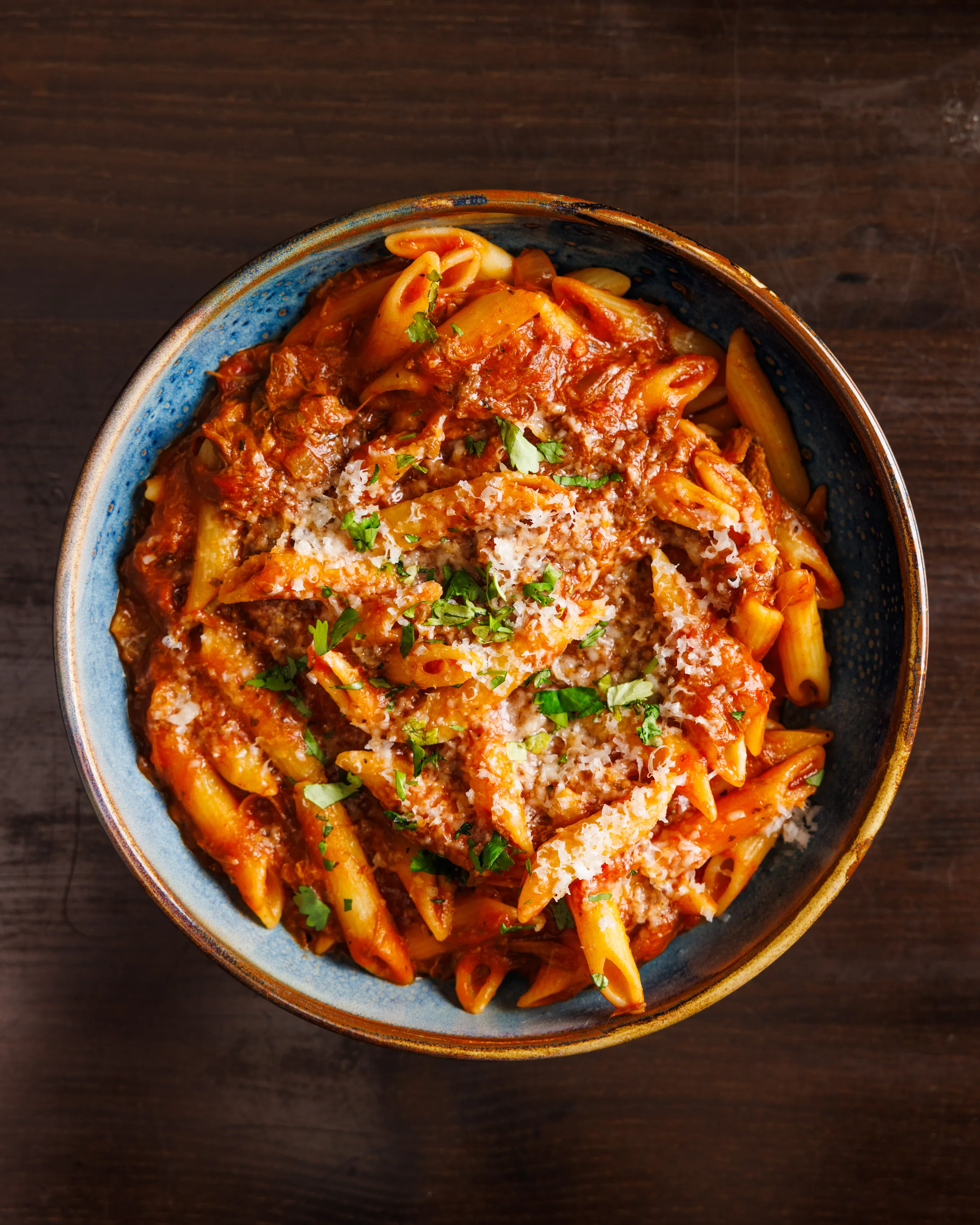 Bowl of penne pasta in rich tomato meat sauce topped with grated cheese and parsley