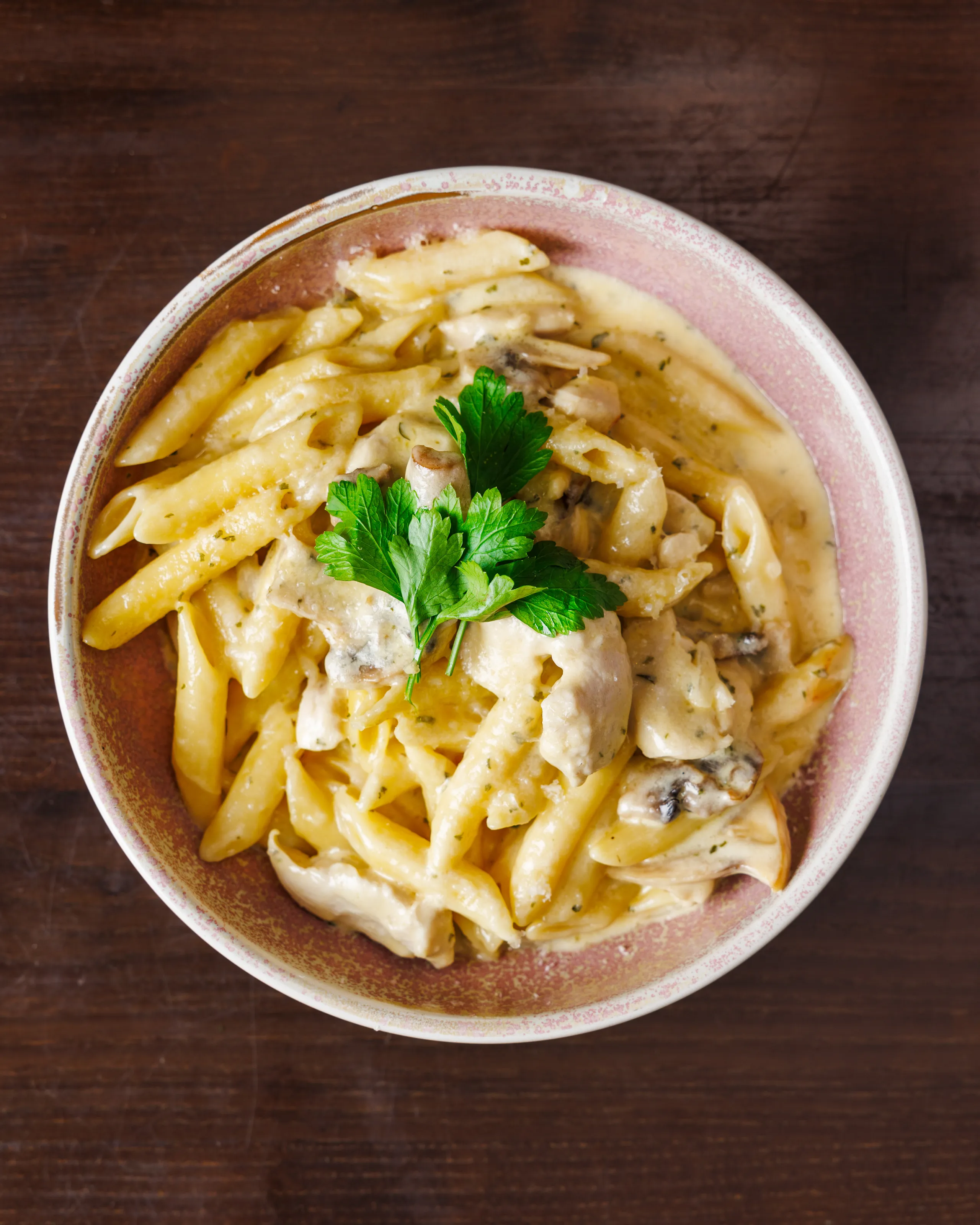 Bowl of creamy penne pasta with mushrooms and parsley garnish on a wooden table
