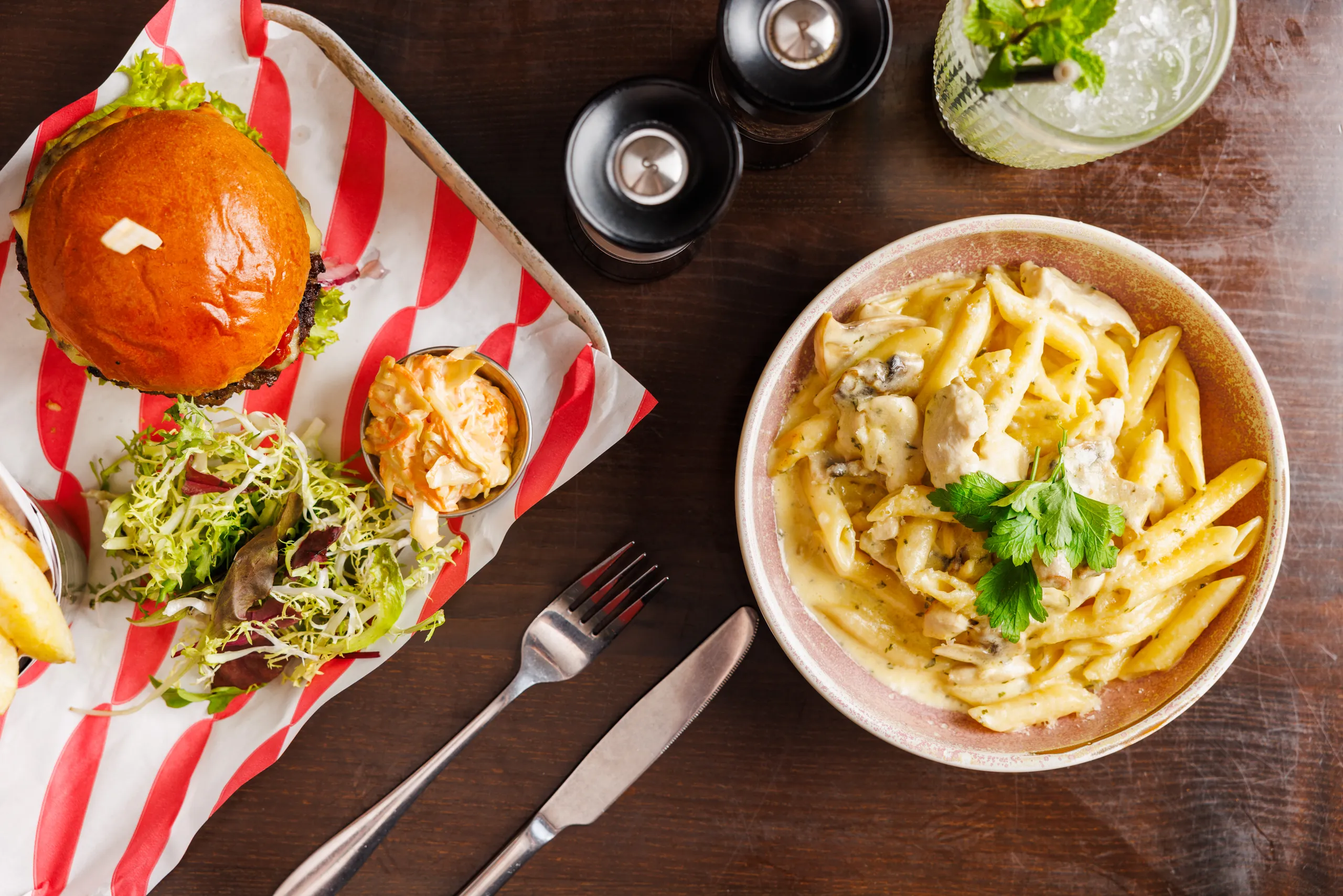 Overhead view of a burger with salad and coleslaw beside creamy chicken pasta on a wooden table.