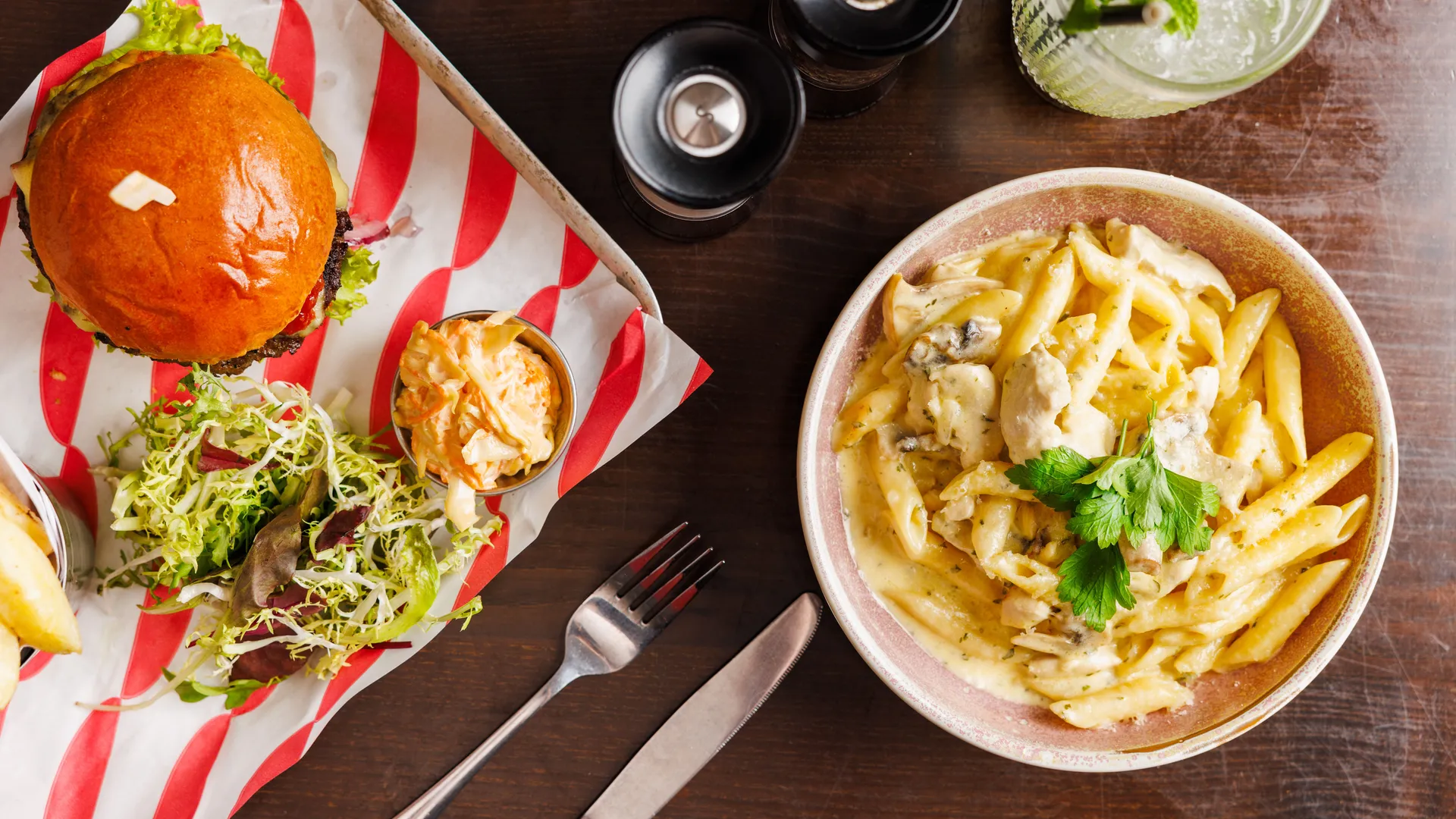 Overhead view of a burger with salad and coleslaw beside creamy chicken pasta on a wooden table.