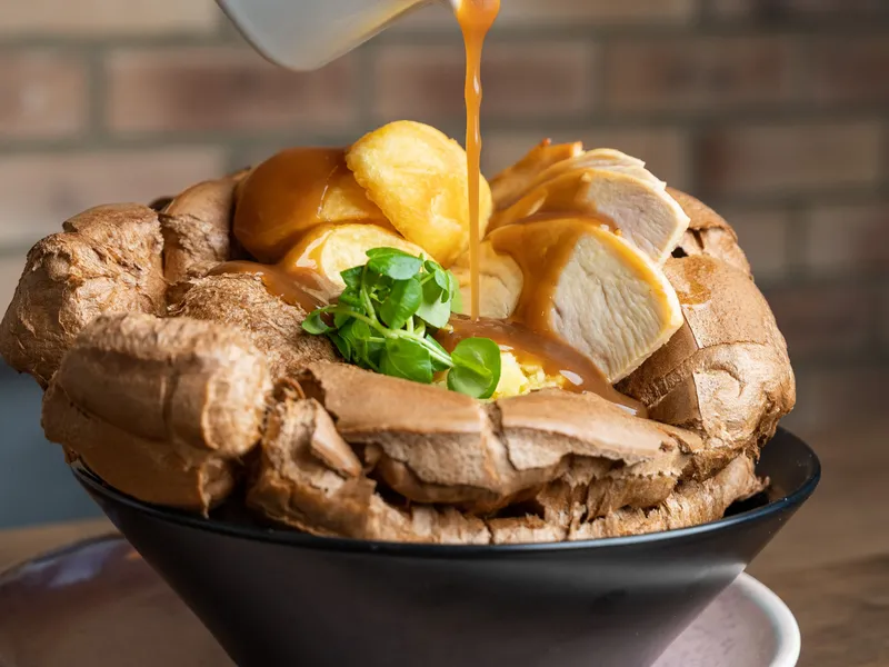 Gravy being poured over roast chicken, Yorkshire pudding and vegetables in a large Yorkshire pudding bowl.