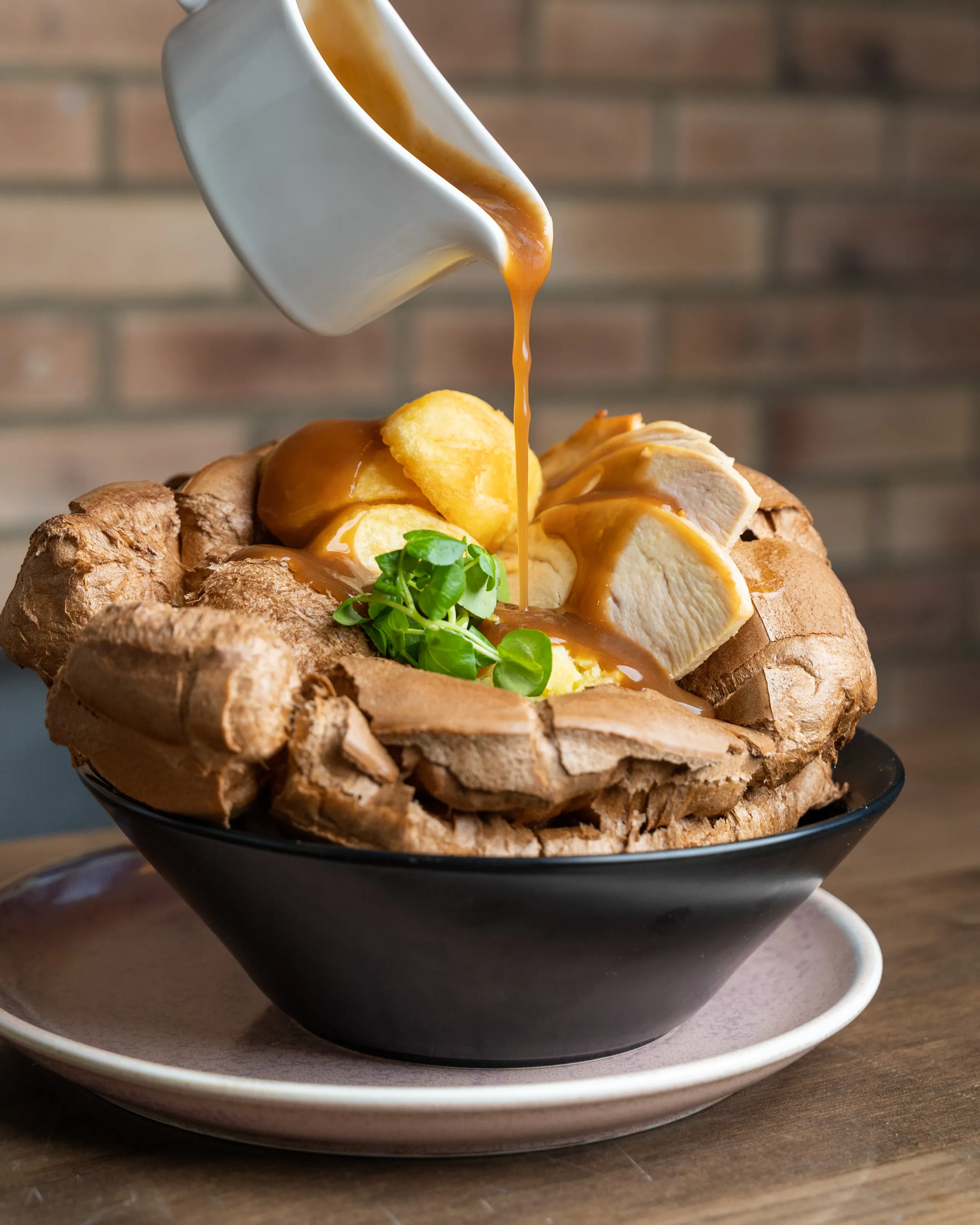 Gravy being poured over roast chicken, Yorkshire pudding and vegetables in a large Yorkshire pudding bowl.