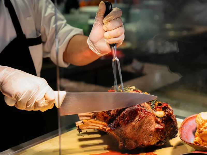 Chef carving a roasted bone-in joint of meat behind a buffet counter