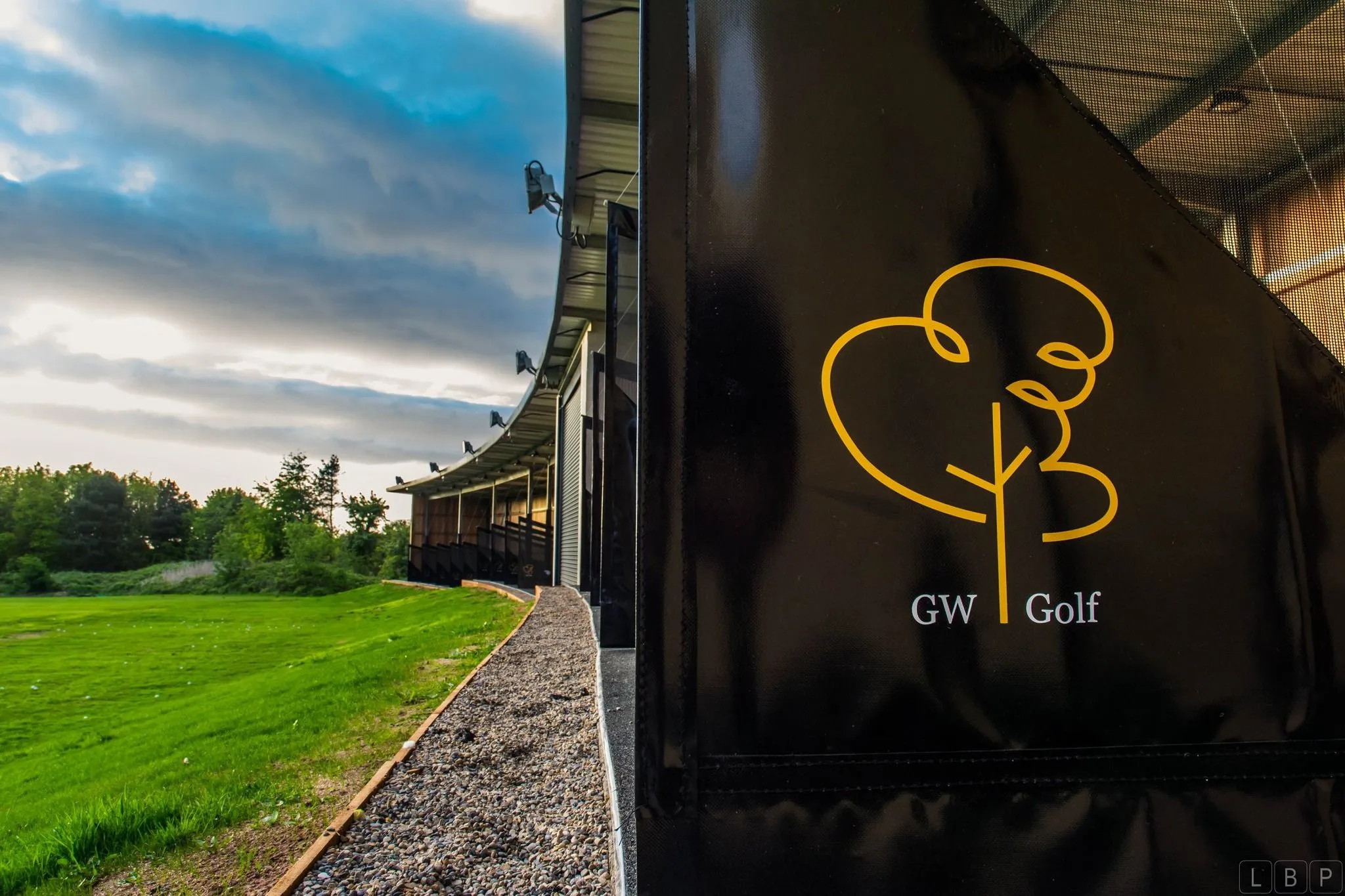 GW Golf driving range building beside a green field beneath a dramatic cloudy sky.