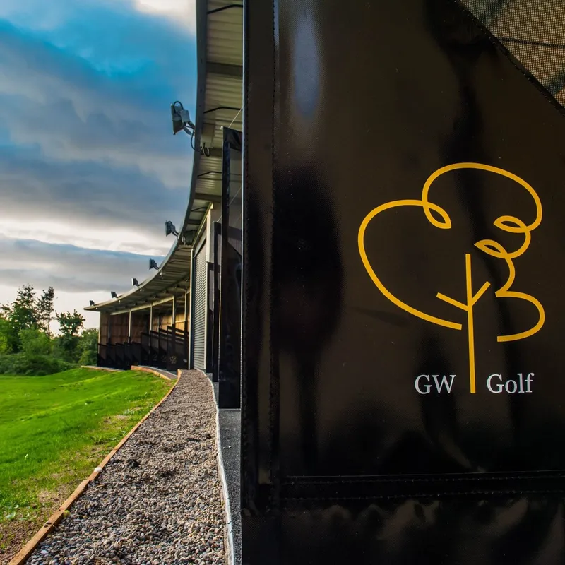 GW Golf driving range building beside a green field beneath a dramatic cloudy sky.
