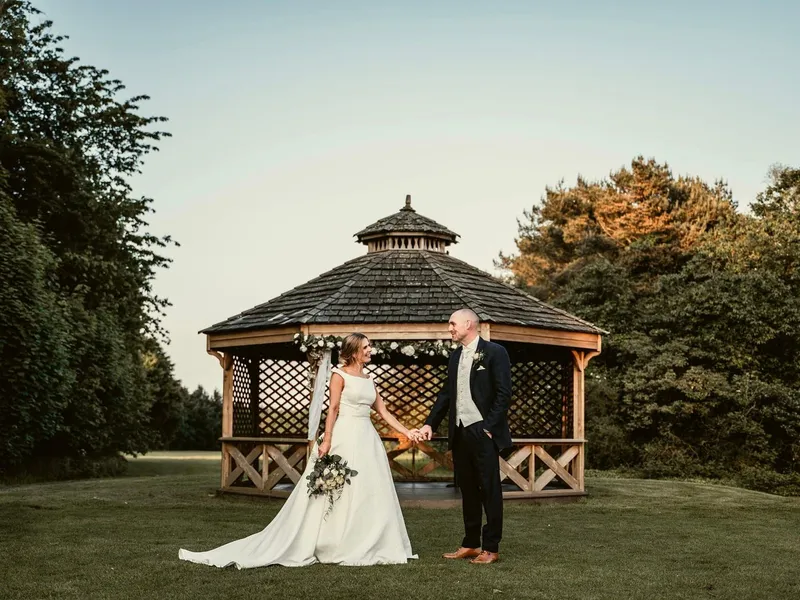 Bride and groom holding hands in front of a flower-decorated wooden gazebo at an outdoor wedding.
