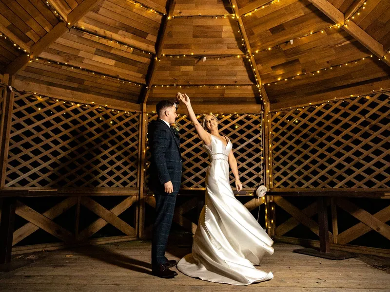 Newlywed couple dancing beneath warm fairy lights in a rustic wooden gazebo at night.