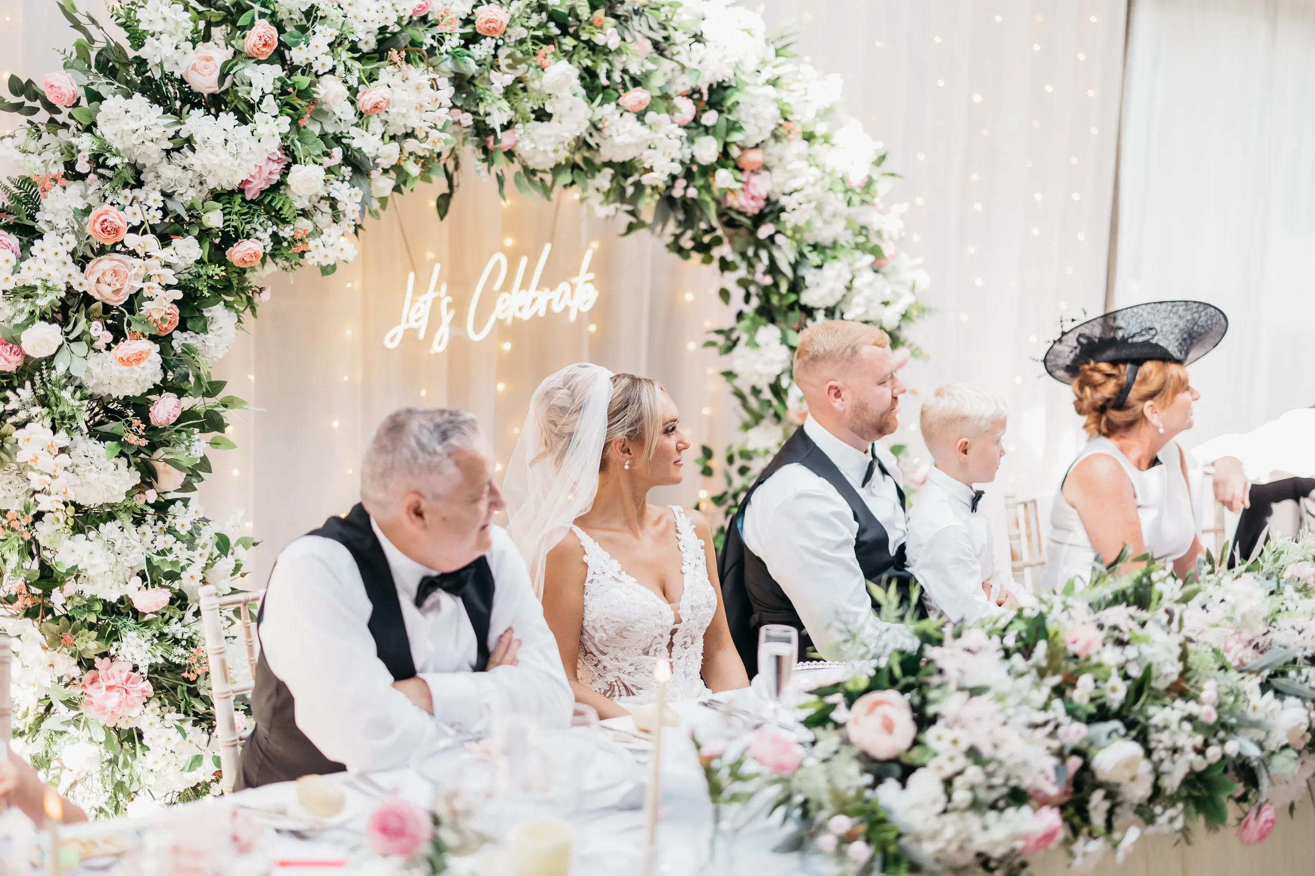 Bride and wedding guests seated beneath a pink and white floral arch at a reception table