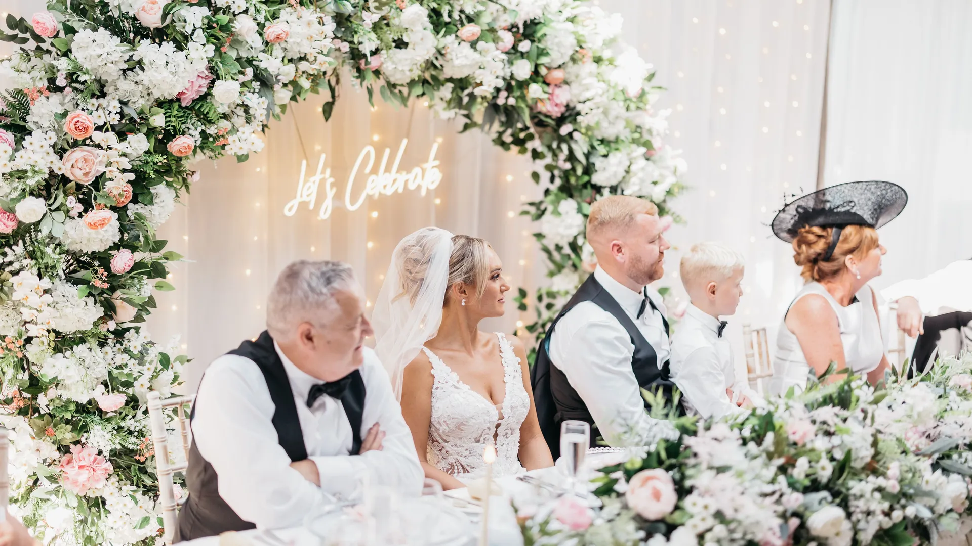 Bride and wedding guests seated beneath a pink and white floral arch at a reception table