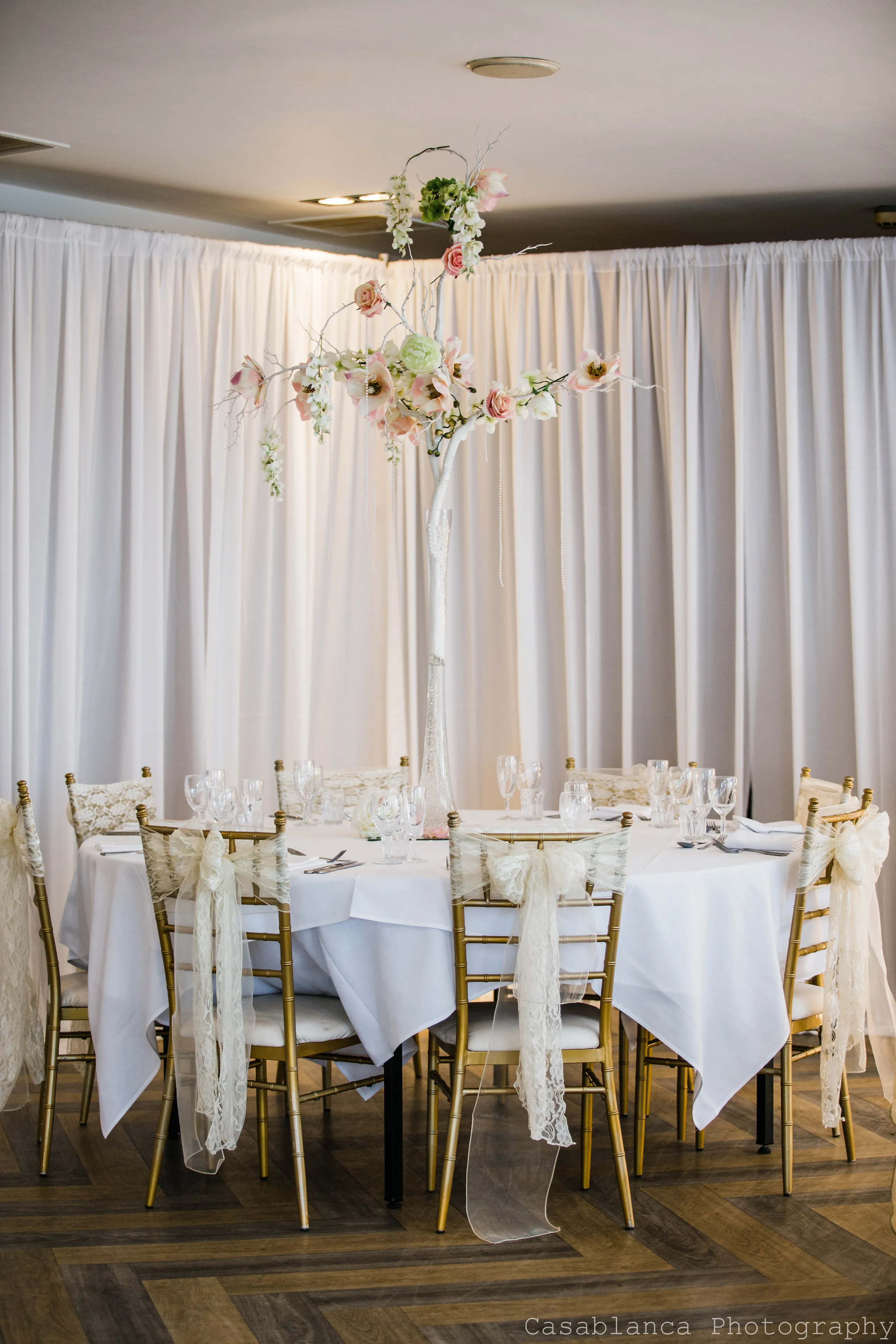 Elegant wedding reception table with a tall floral tree centrepiece, gold chairs and lace bows.