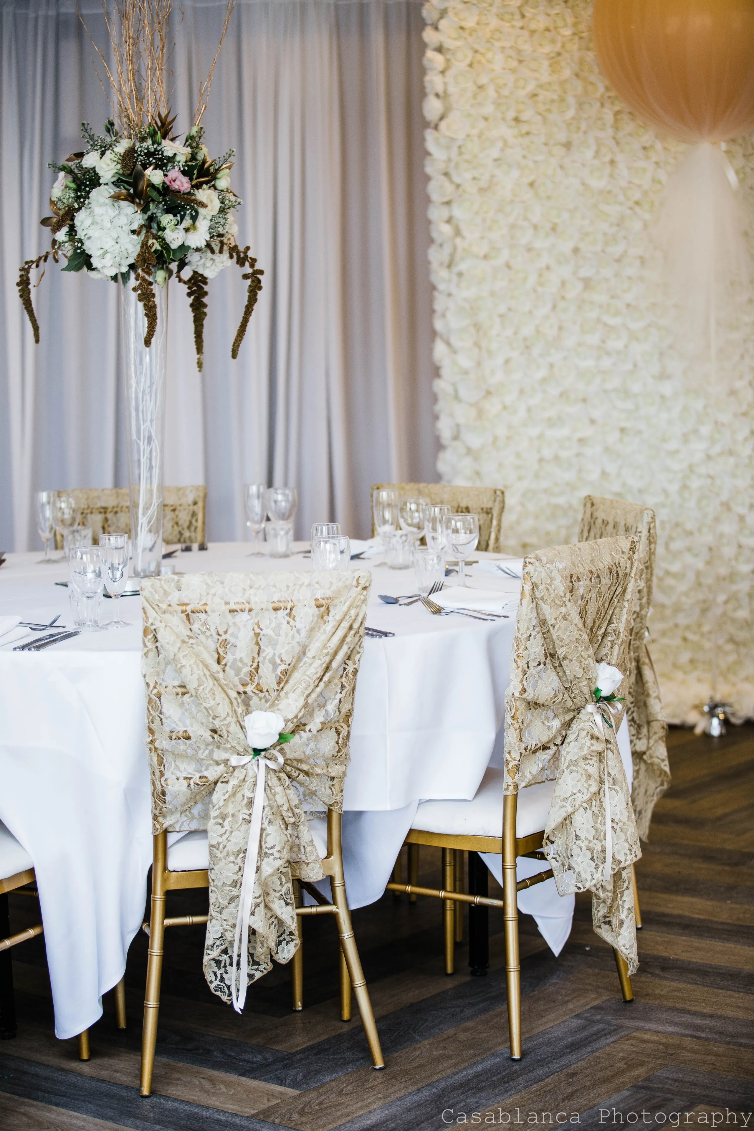 Elegant wedding reception table with lace-covered gold chairs, a tall floral centrepiece and a white rose wall.