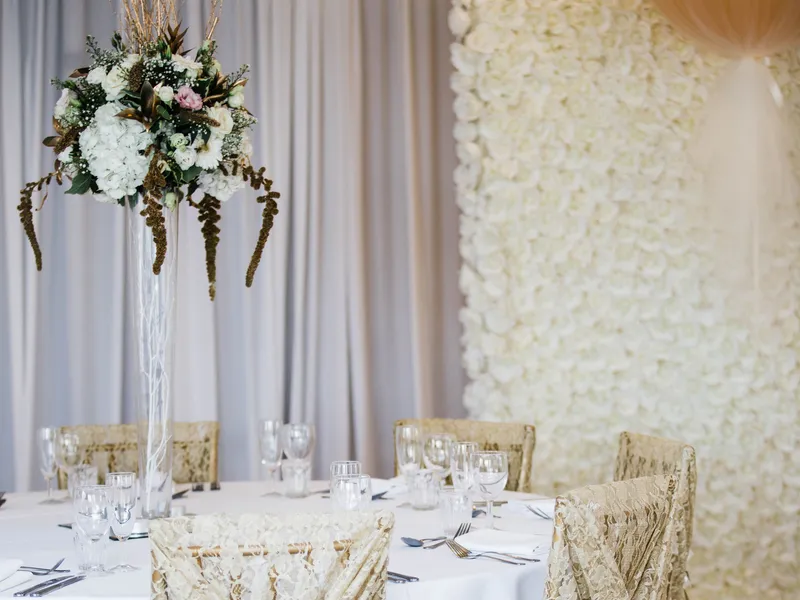 Elegant wedding reception table with lace-covered gold chairs, a tall floral centrepiece and a white rose wall.