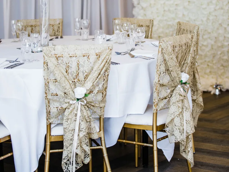Elegant wedding reception table with lace-covered gold chairs, a tall floral centrepiece and a white rose wall.