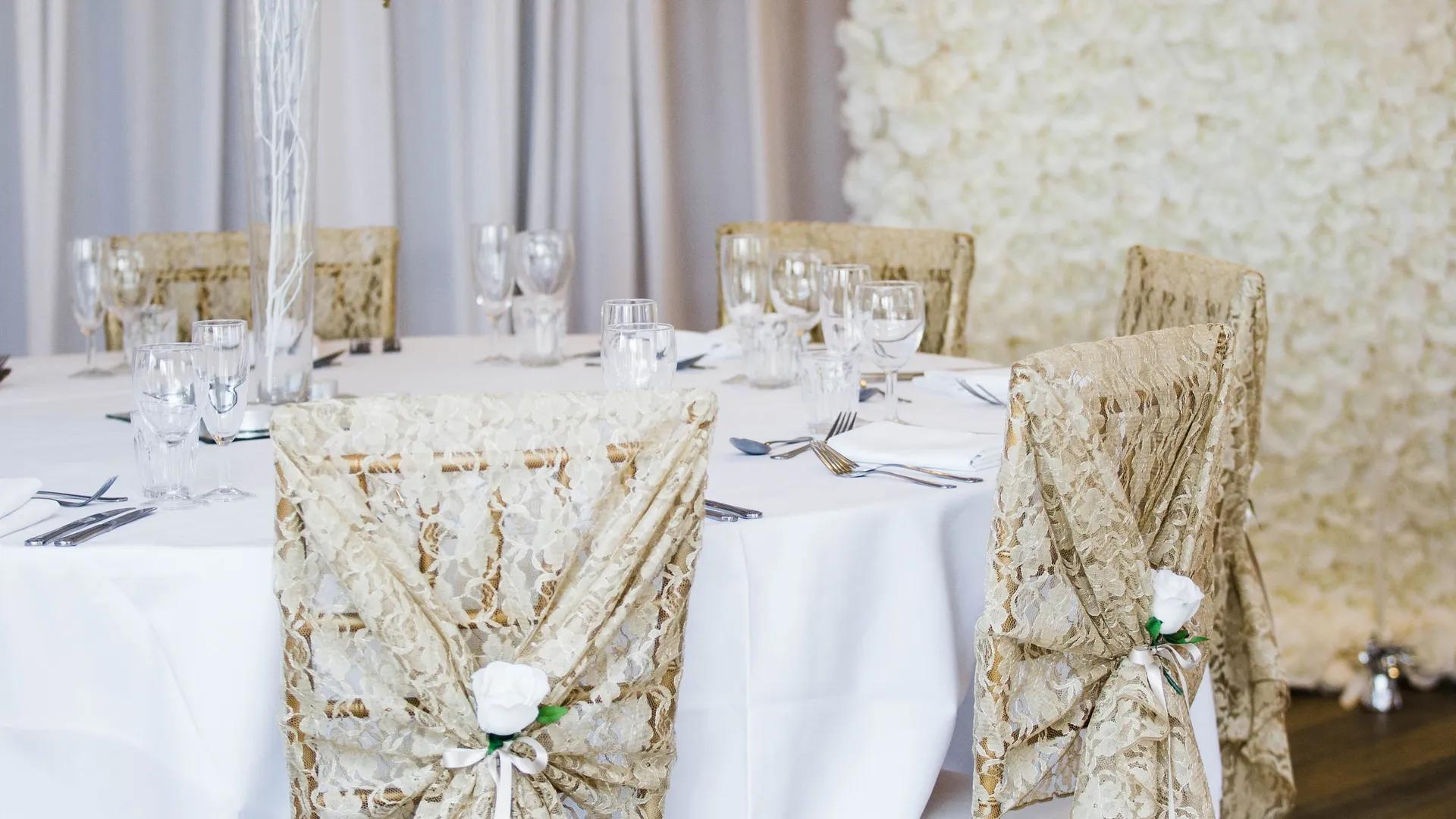 Elegant wedding reception table with lace-covered gold chairs, a tall floral centrepiece and a white rose wall.