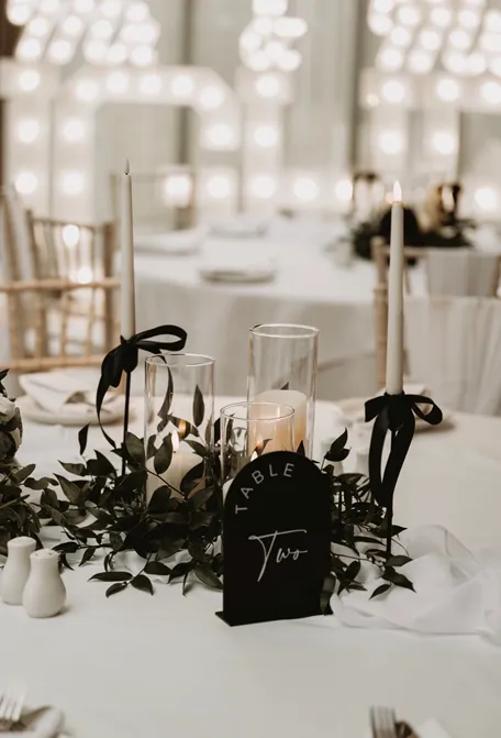 Elegant wedding reception table with black-ribbon candles, greenery, glassware and a table number sign beneath glowing marquee lights.