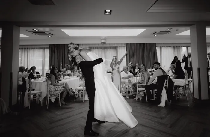 Black-and-white photograph of a groom lifting and kissing his bride during their wedding reception as guests applaud.