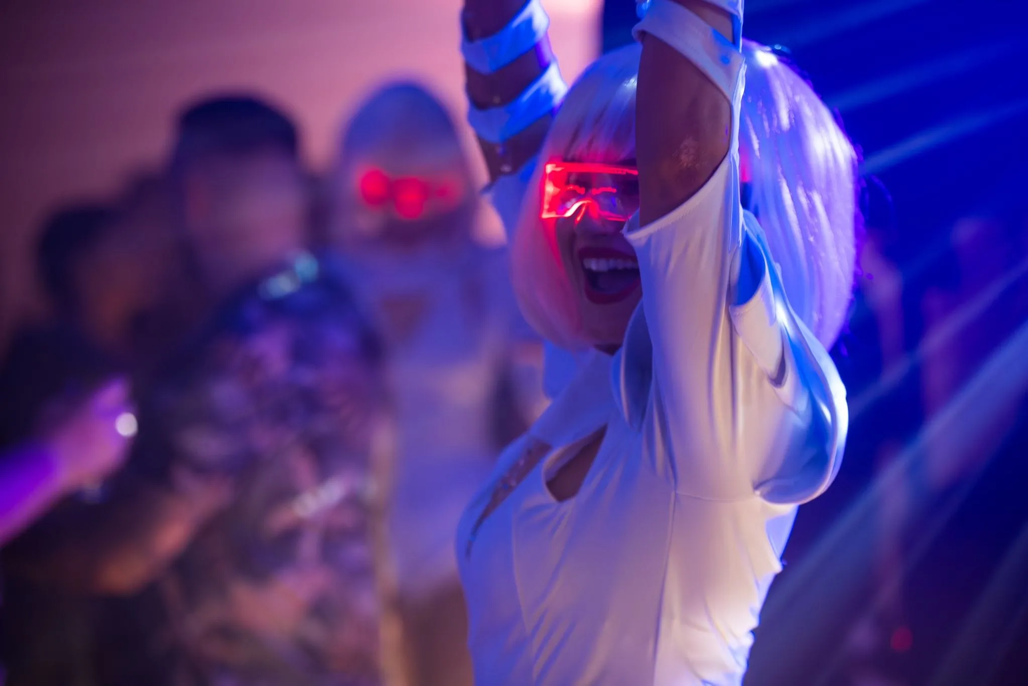 Smiling partygoer in a white outfit and red neon visor dancing under blue and purple lights.