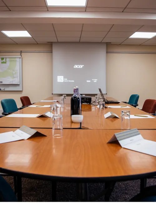 Prepared conference room with a large wooden table, colourful chairs, notebooks, water glasses and a projection screen.