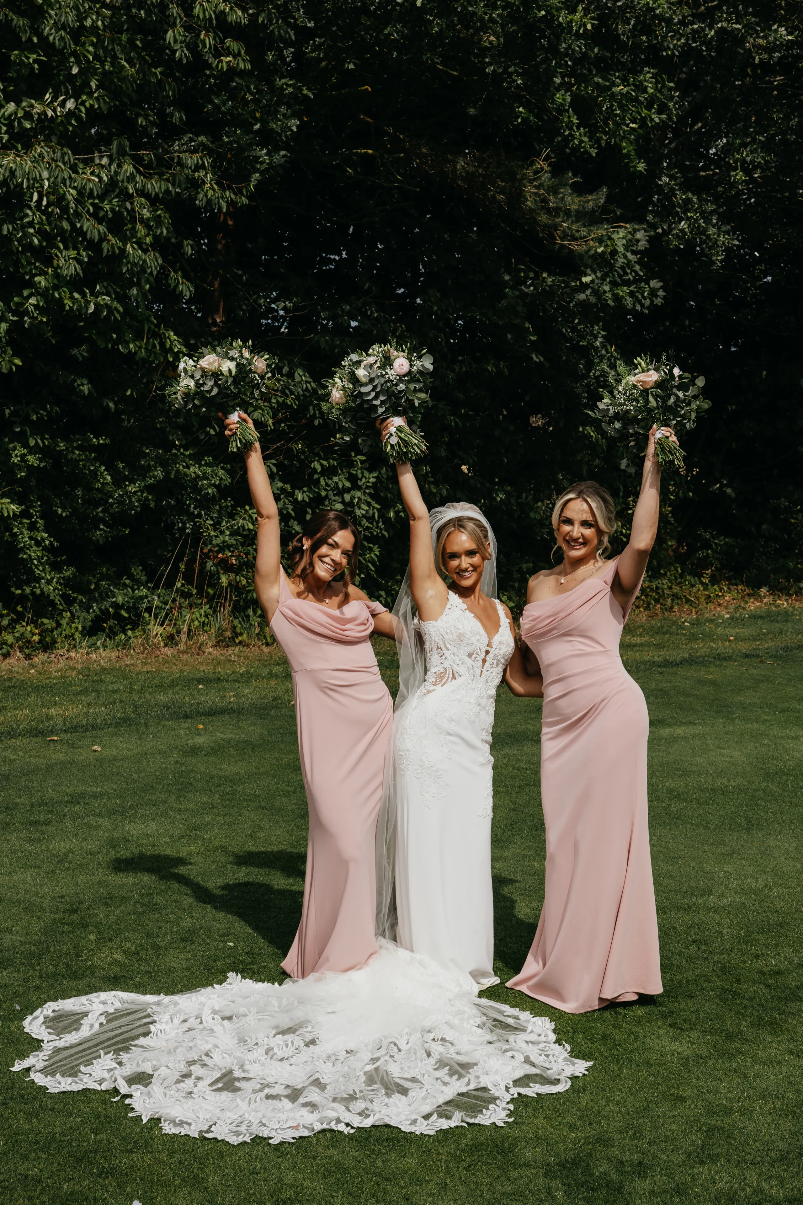 Bride in a white lace wedding gown with two bridesmaids in blush pink dresses raising bouquets on a lawn