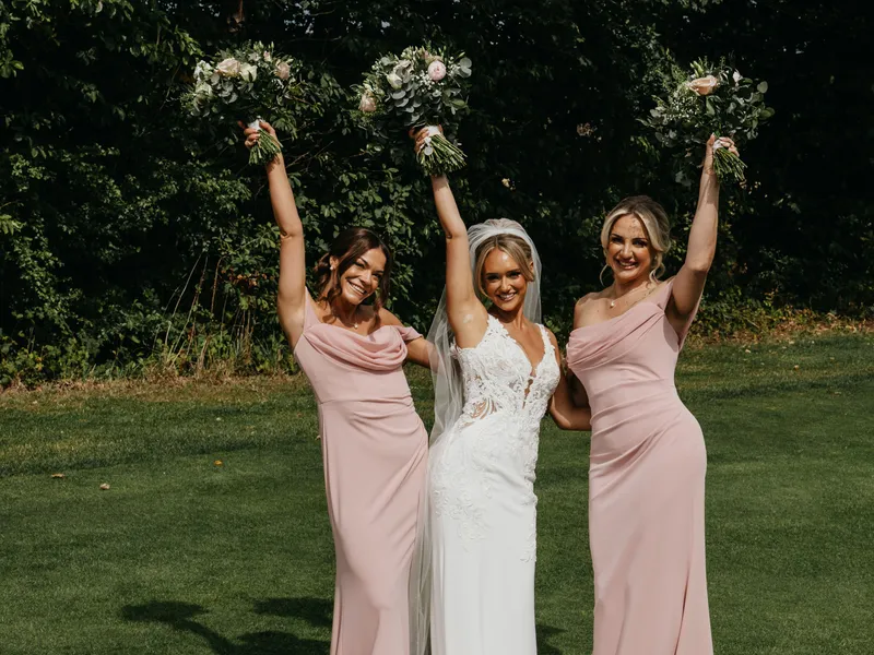 Bride in a white lace wedding gown with two bridesmaids in blush pink dresses raising bouquets on a lawn