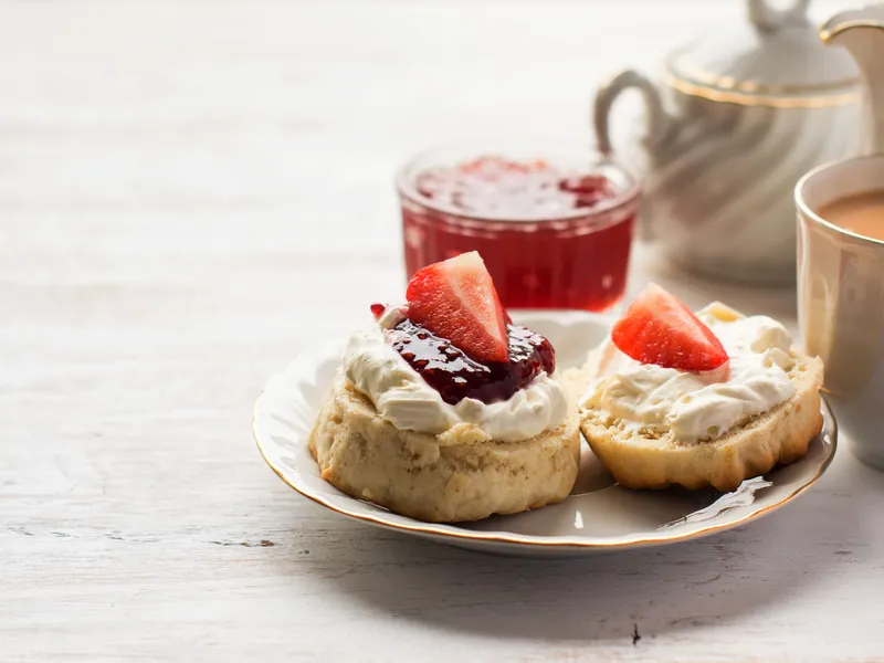 Two cream scones topped with strawberry jam and fresh strawberries beside a teapot and cup.