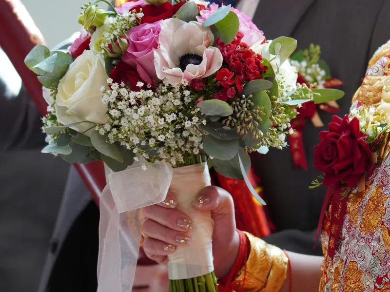 Bride in ornate red and gold traditional attire holding a colourful wedding bouquet beside a groom in a dark suit.