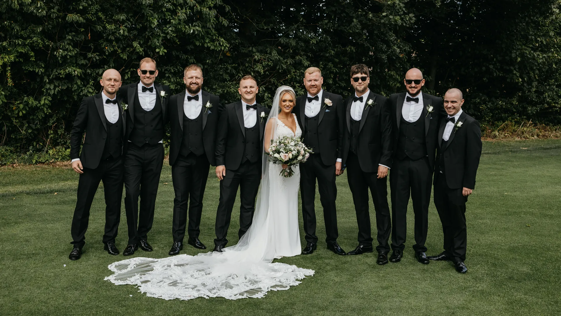 Bride and groom standing with groomsmen in black tuxedos on a lawn