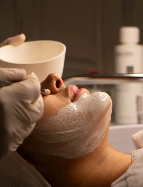 Client receiving a facial treatment as a gloved beautician applies product with a bowl nearby.