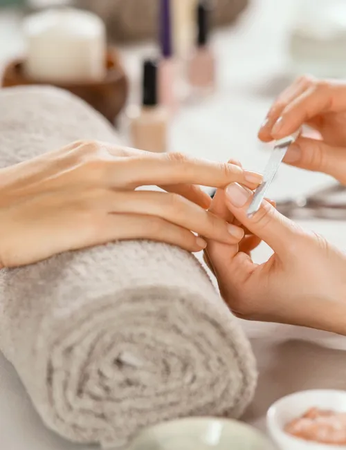 Close-up of a manicure technician filing a client's fingernail beside a rolled grey towel.