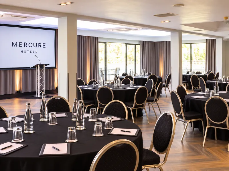 Hotel conference room arranged with black-clothed tables, chairs, water bottles and a Mercure Hotels presentation screen.