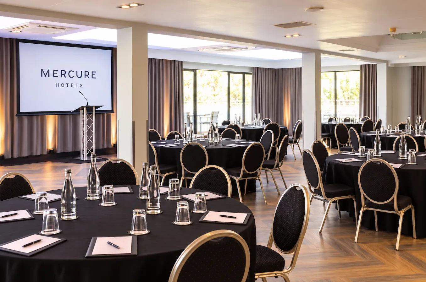 Hotel conference room arranged with black-clothed tables, chairs, water bottles and a Mercure Hotels presentation screen.