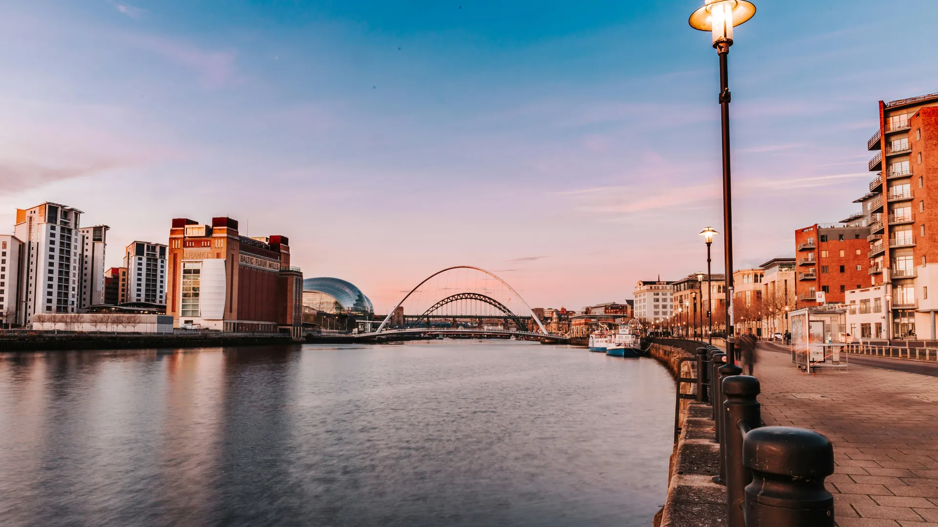 The River Tyne at dusk, with the Gateshead Millennium Bridge, Baltic Centre and Newcastle Quayside beneath a pink and blue sky.