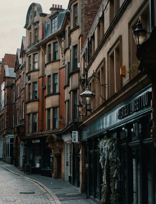 Empty cobbled street lined with historic brick and stone buildings, shopfronts and ornate street lamps.