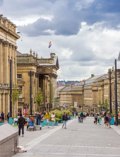 Pedestrians on a broad Newcastle city centre street lined with historic stone buildings, with a rainbow flag flying beneath a cloudy sky.