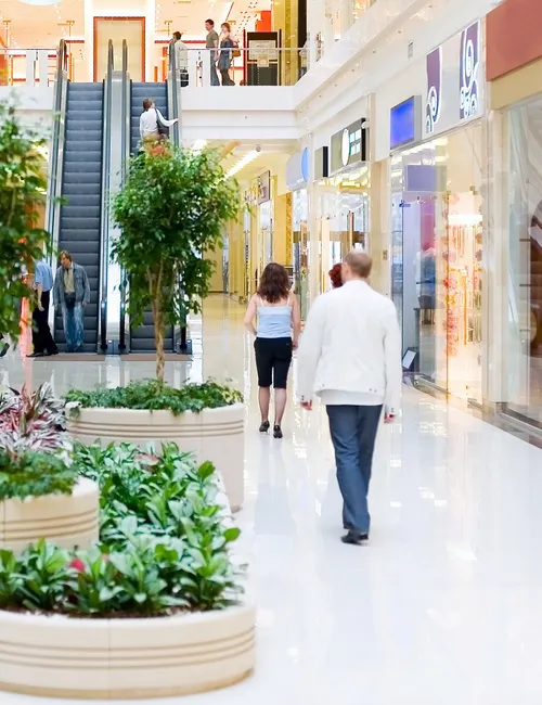 Shoppers walking through a bright indoor shopping centre with shopfronts, escalators and large planters