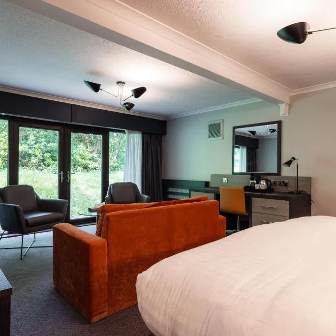 Modern hotel room with a large bed, orange sofa, armchair, desk and garden-facing windows.