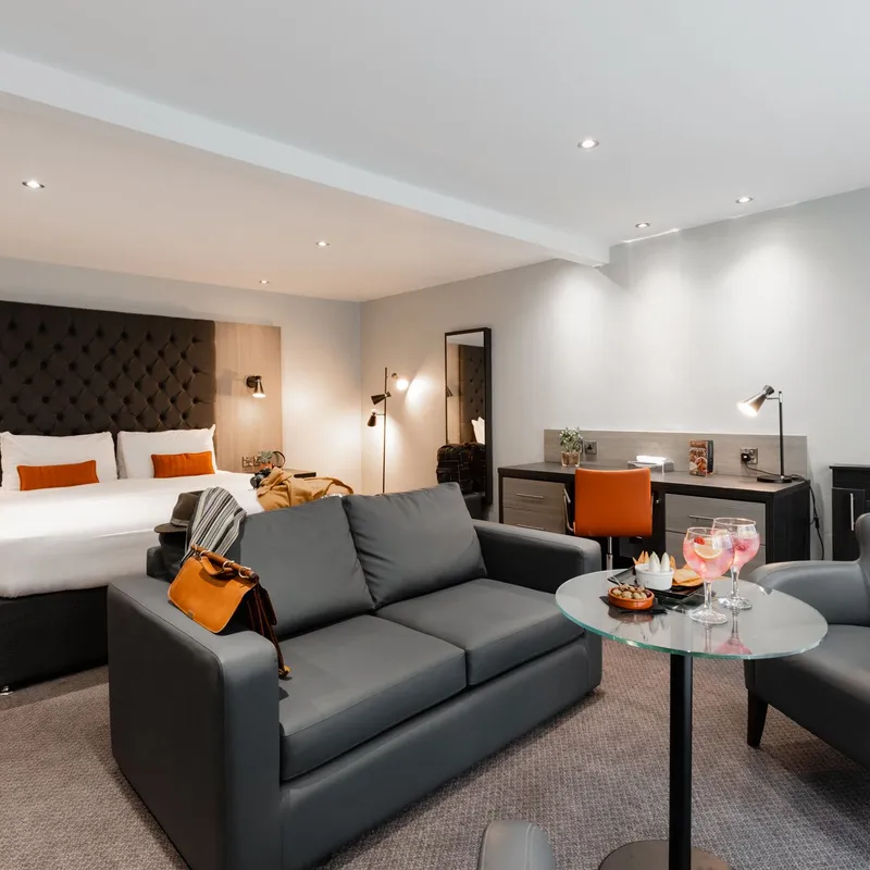 Modern grey hotel suite with a double bed, sofa, armchair, desk and glass coffee table.