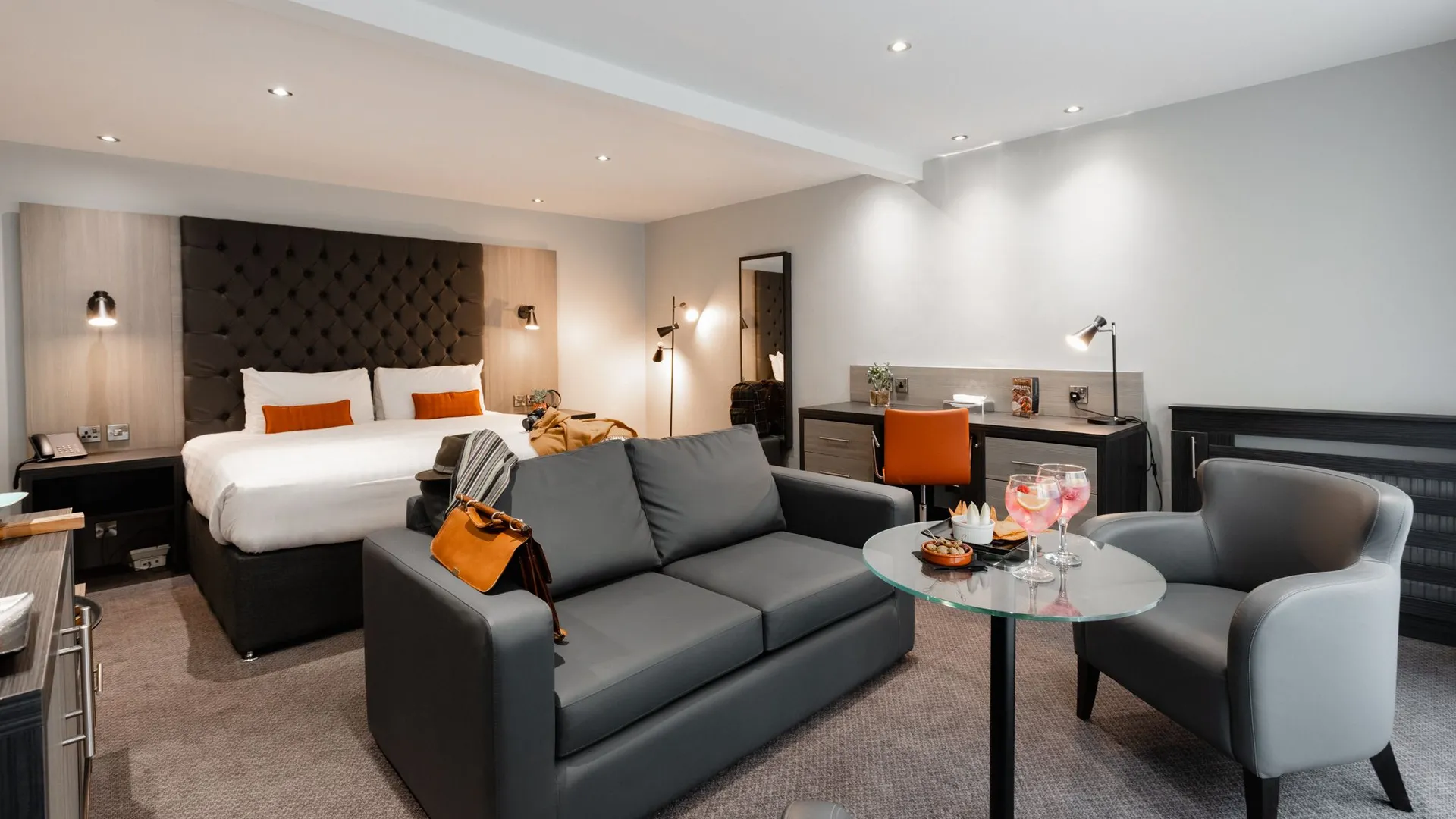 Modern grey hotel suite with a double bed, sofa, armchair, desk and glass coffee table.