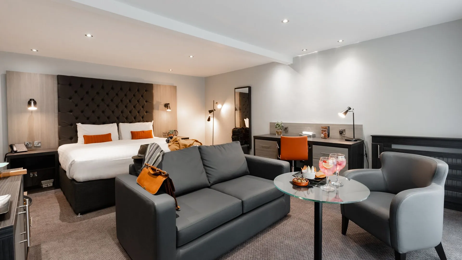 Modern grey hotel suite with a double bed, sofa, armchair, desk and glass coffee table.