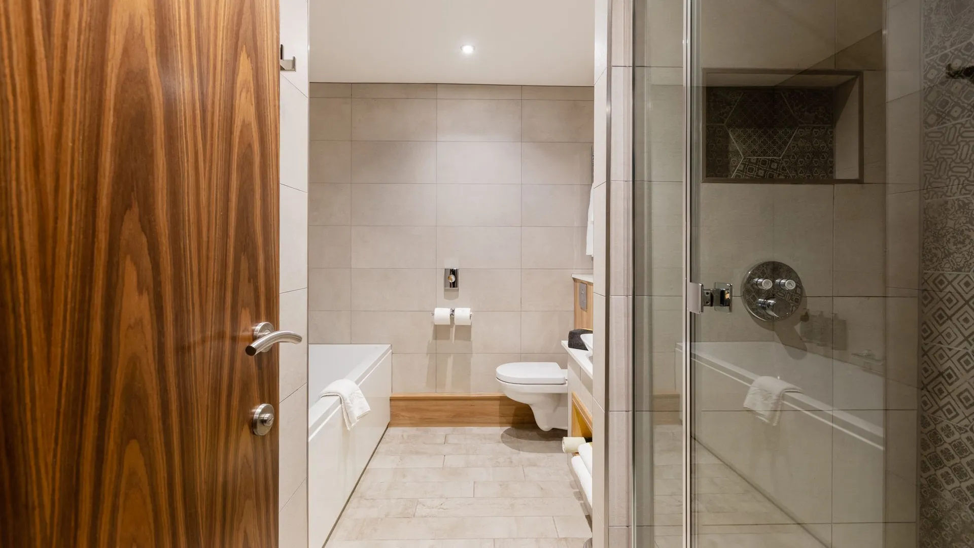 Modern bathroom with a wooden door, bathtub, wall-mounted toilet and glass shower enclosure.