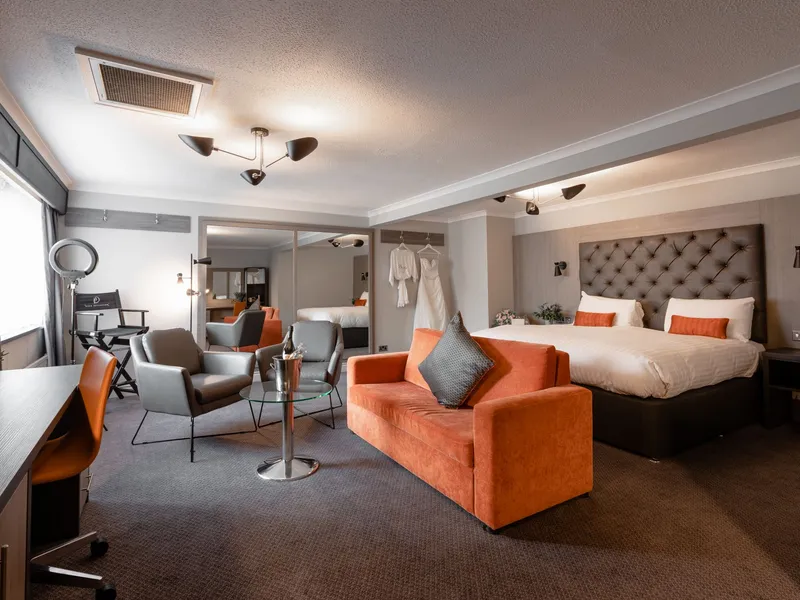 Spacious hotel bridal suite with a king-size bed, orange armchair, dressing area and wedding dress.