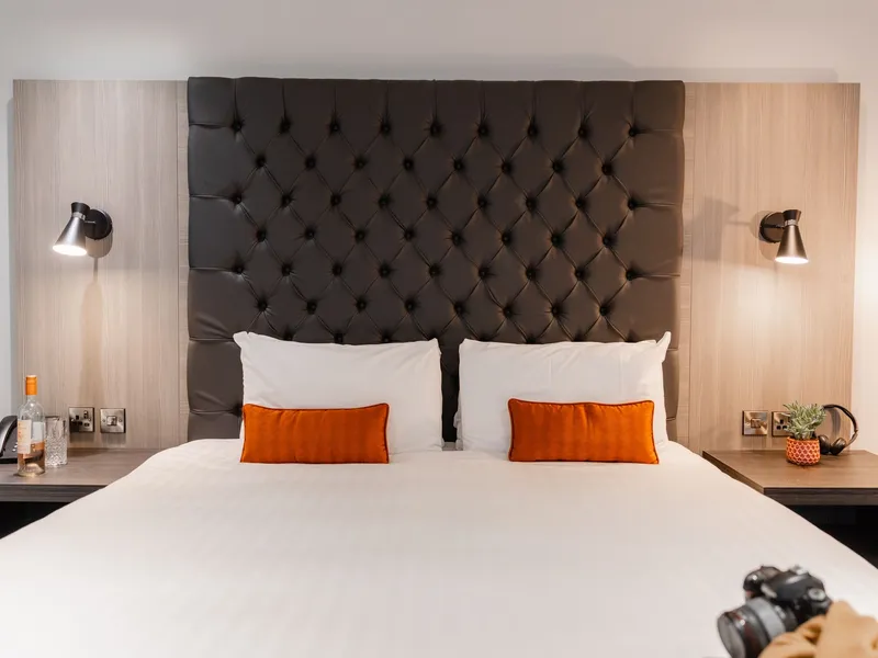 Modern hotel bedroom with a dark tufted headboard, white bedding and orange cushions.