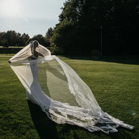 Bride in a white wedding dress holding a long lace veil across a sunlit lawn beside tall trees.