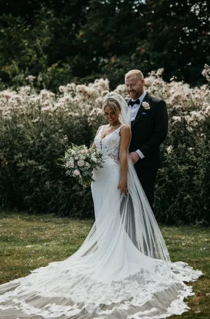 Bride in a lace wedding gown with a long train and bouquet standing with groom in a garden