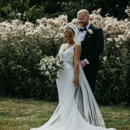 Bride in a lace wedding gown with a long train and bouquet standing with groom in a garden